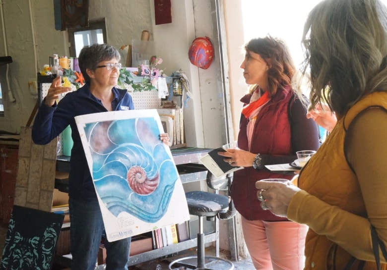 Abby Hogan, Scenic Charge Artist is holding up a painting explaining to two other people how a floor stage design comes together